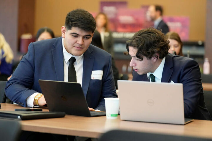 Two male graduate students look at laptops in a classroom.